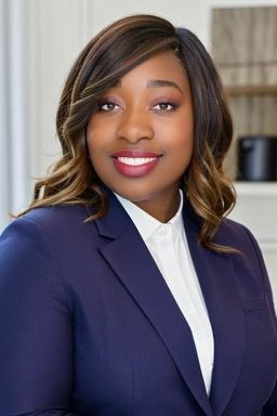 Person wearing a navy blue blazer and white shirt, standing indoors with a neutral background.