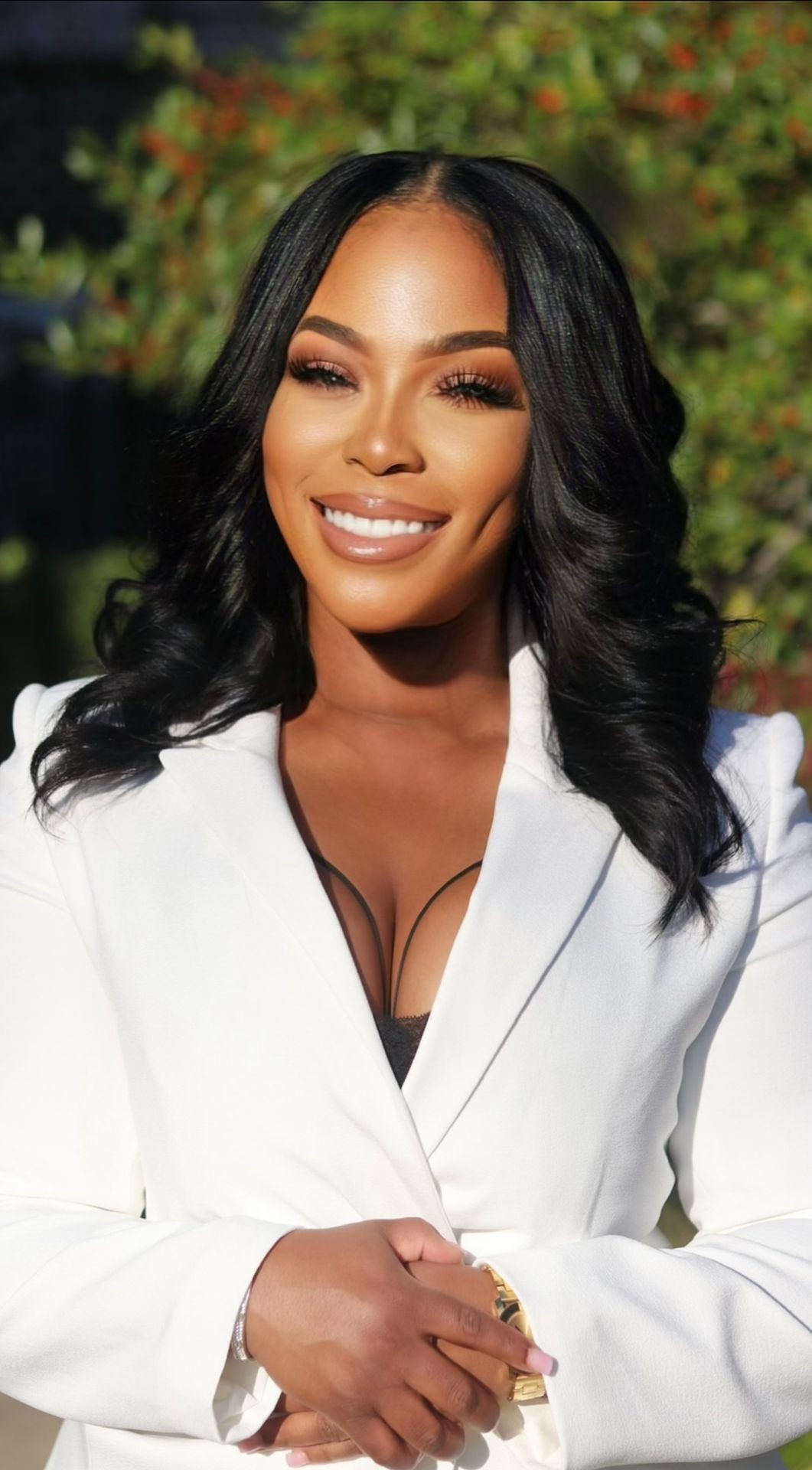 Woman smiling and wearing a white blazer, with long black hair in a natural outdoor setting.