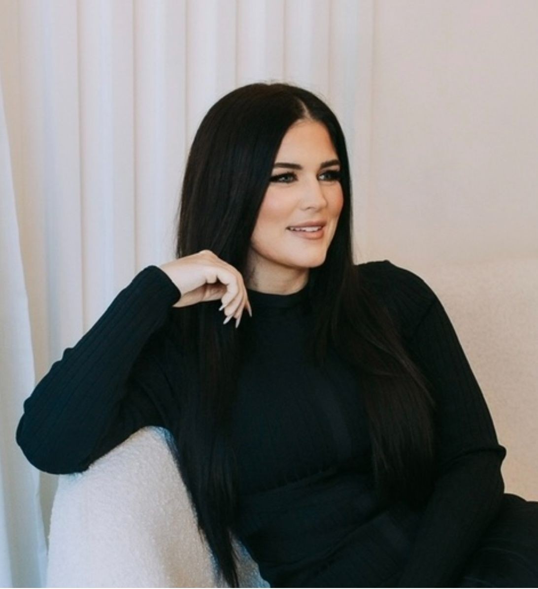 Woman with long dark hair wearing a black outfit, sitting on a white chair and smiling.