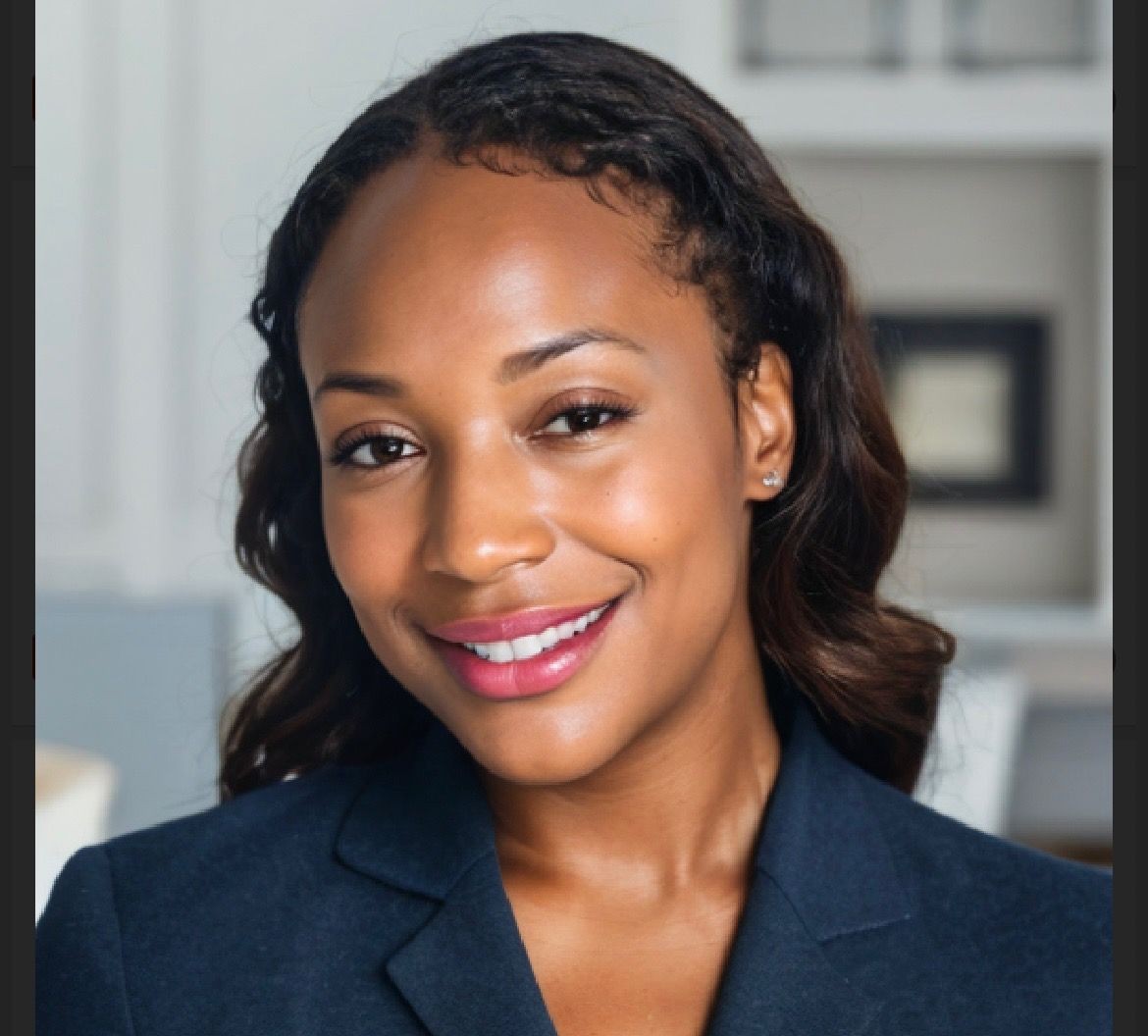 Woman in a dark blazer smiling, with a blurred background of indoor setting.