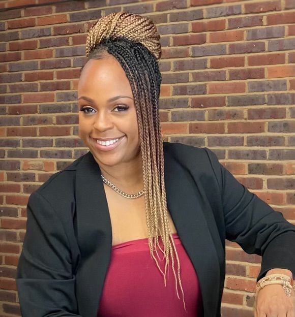 Person wearing a black blazer and red top, posing against a brick wall background.